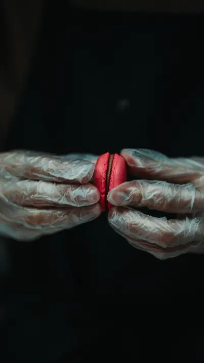 A pink macaron held between two gloved hands against black