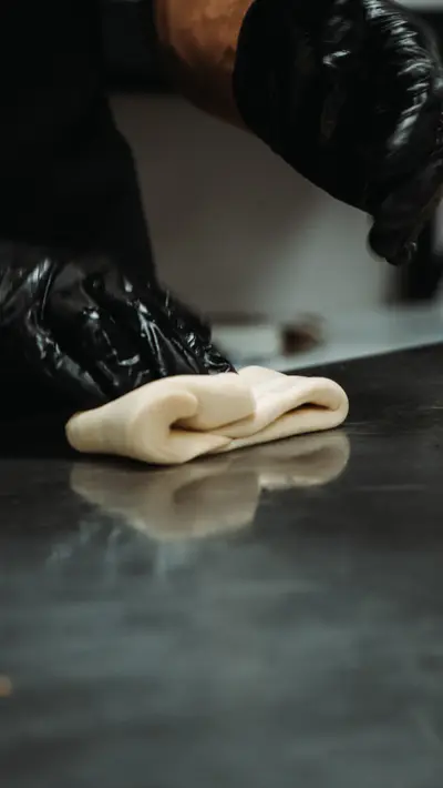 Gloved hands folding dough on a steel bench