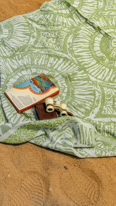 A green printed towel on sand with a book and sunglasses