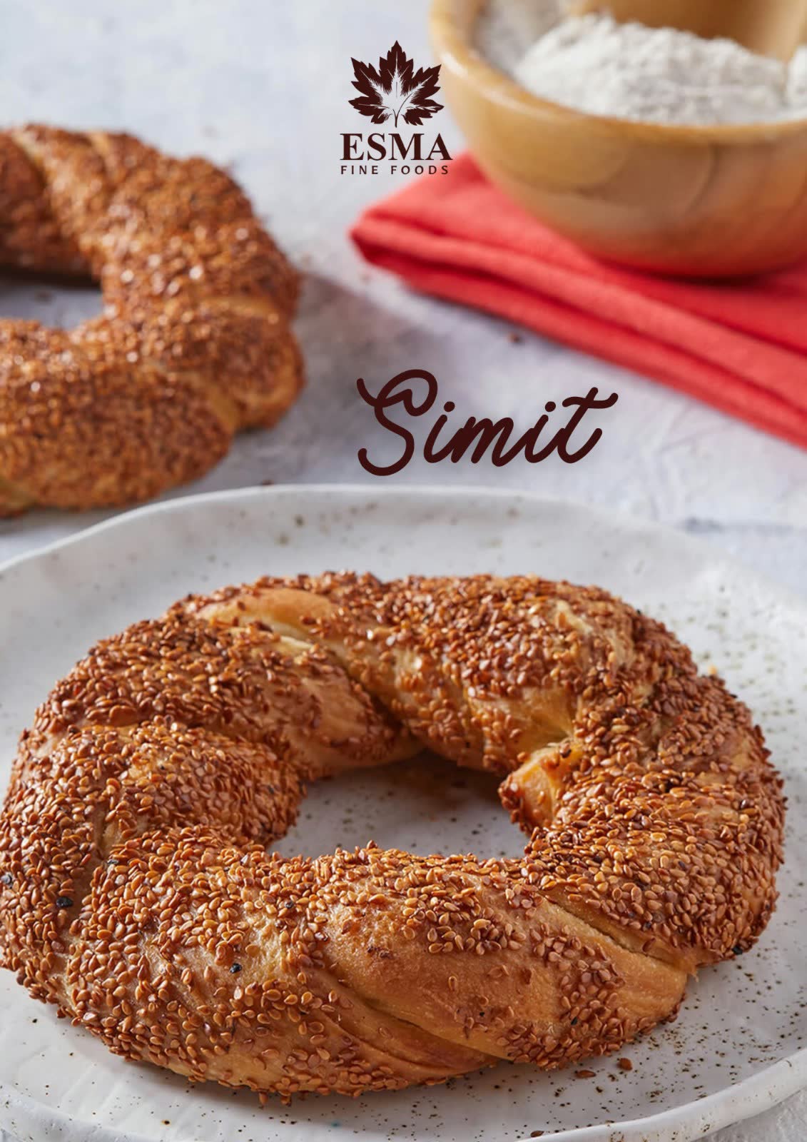 Sesame-crusted simit on a white plate beside a bowl of flour and a coral napkin
